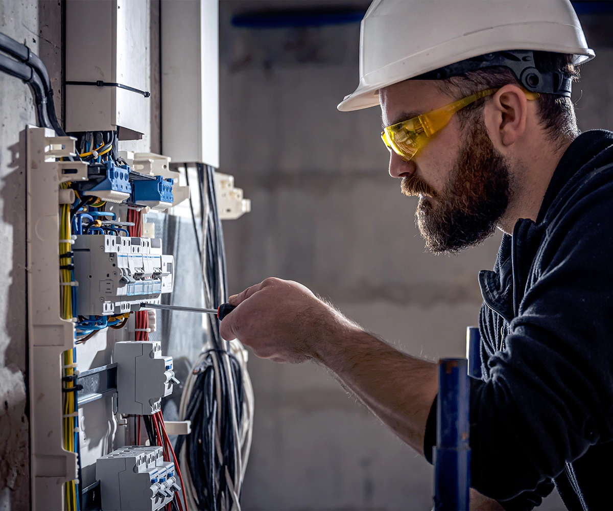 A man in white hard hat and safety glasses working on electrical equipment.